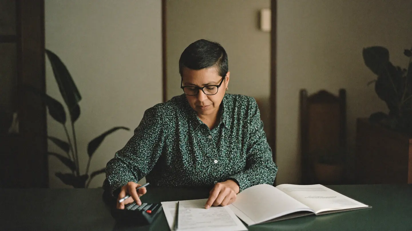 Mulher de óculos confere documentos e faz contas na calculadora na mesa de casa